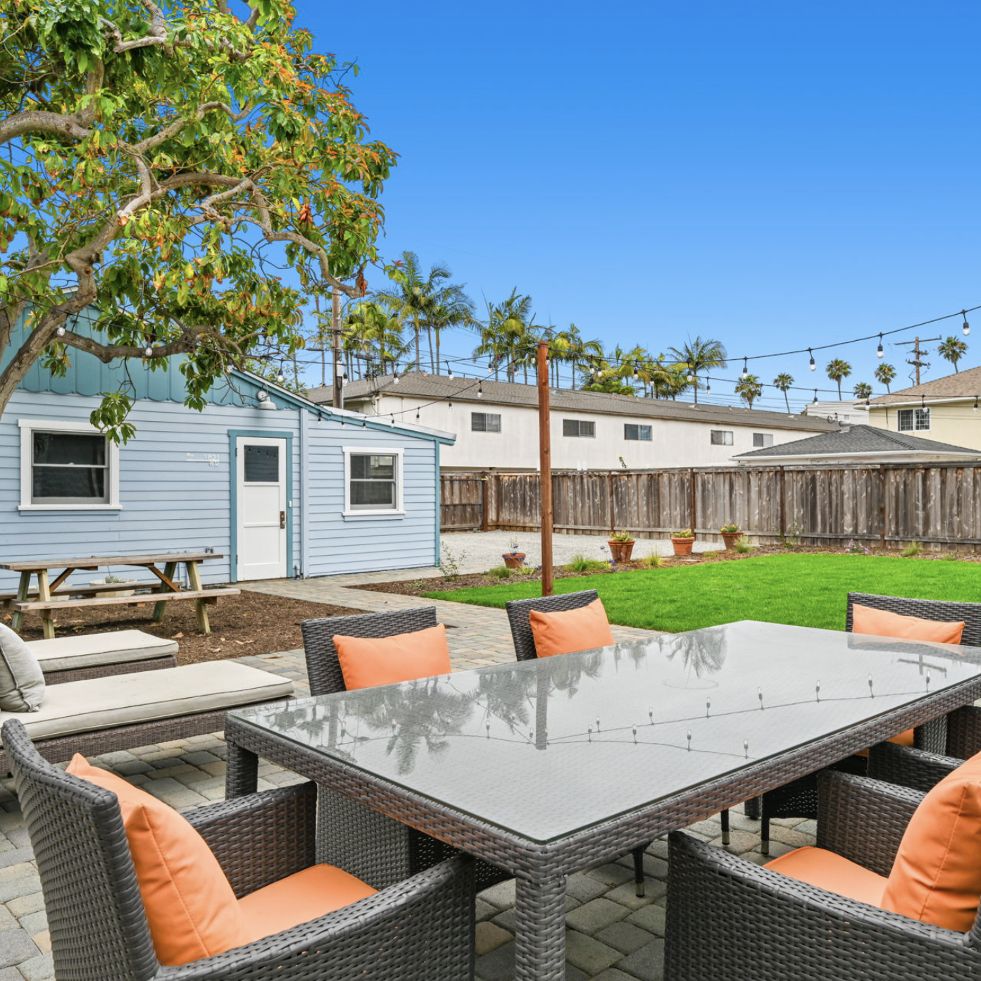 Renovated backyard patio featuring stone pavers, an outdoor dining set with orange cushions, and a blue home exterior with string lights.